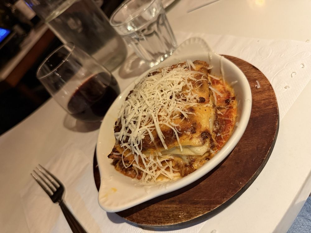 A close-up of baked lasagna topped with grated cheese in a white oval dish on a wooden board, with a glass of red wine and a glass of water in the background on a set dining table.
