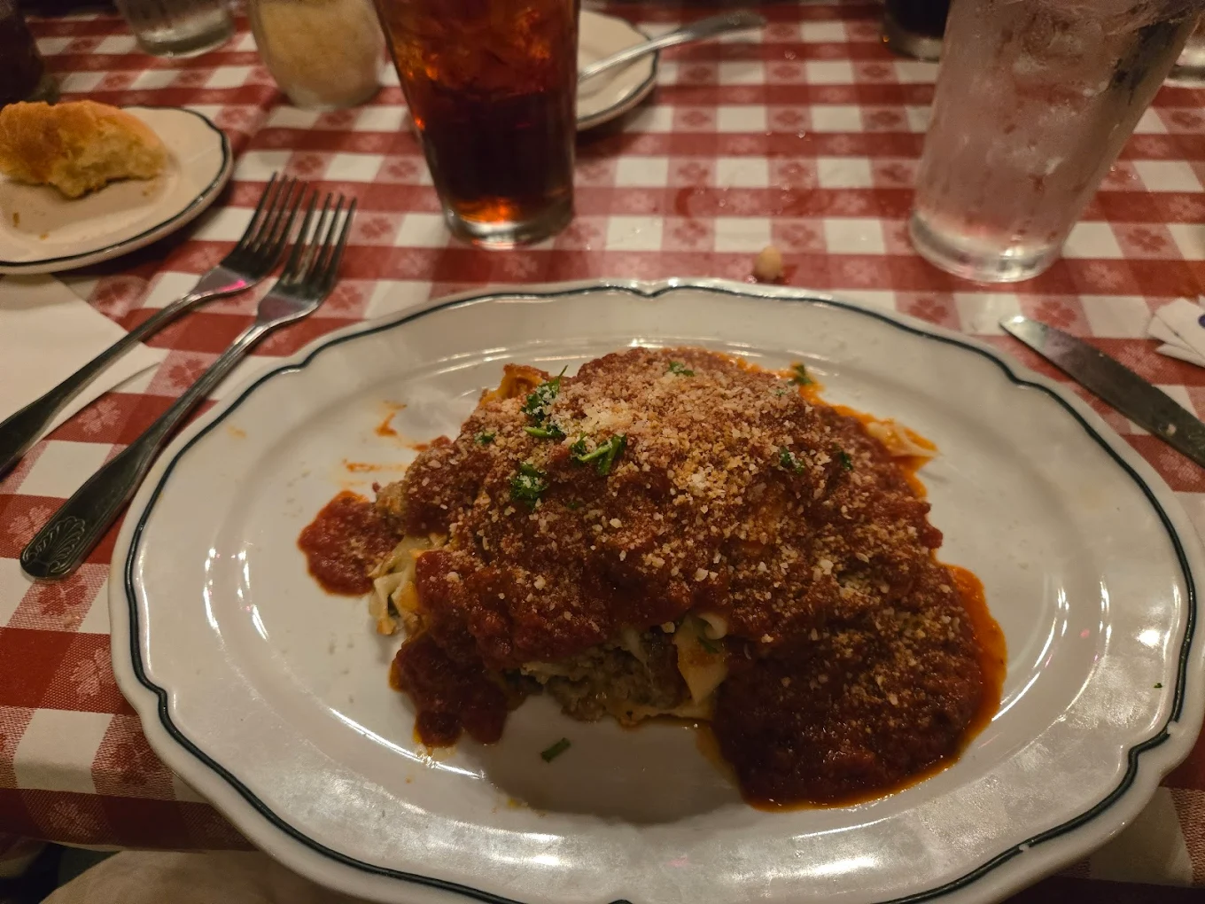 A plate of lasagna topped with marinara sauce and grated cheese sits on a white plate, on a red-and-white checkered tablecloth, with a glass of iced tea and utensils nearby.