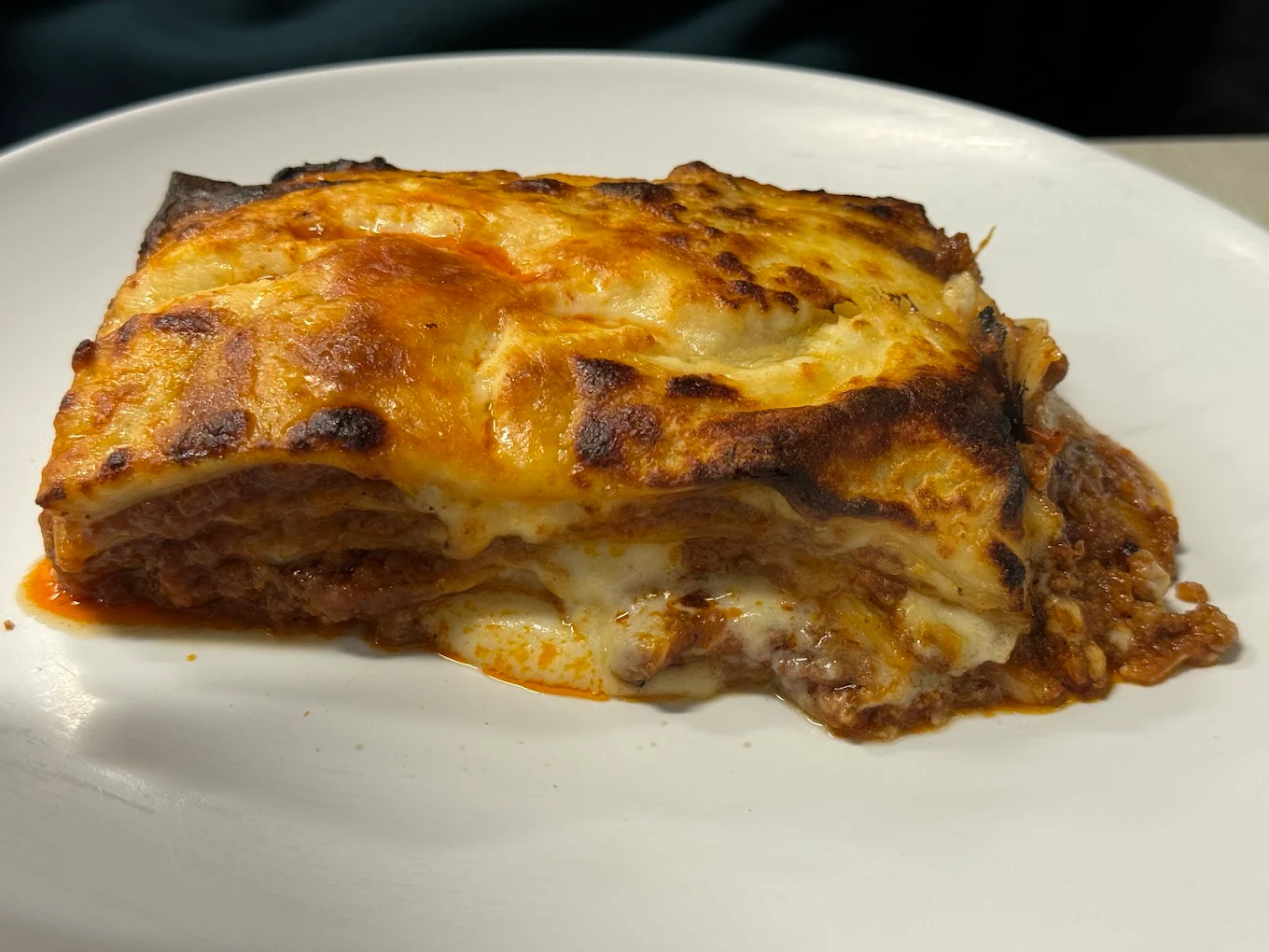 A close-up of a slice of lasagna on a white plate, showing layers of pasta, meat sauce, and melted cheese browned on top.