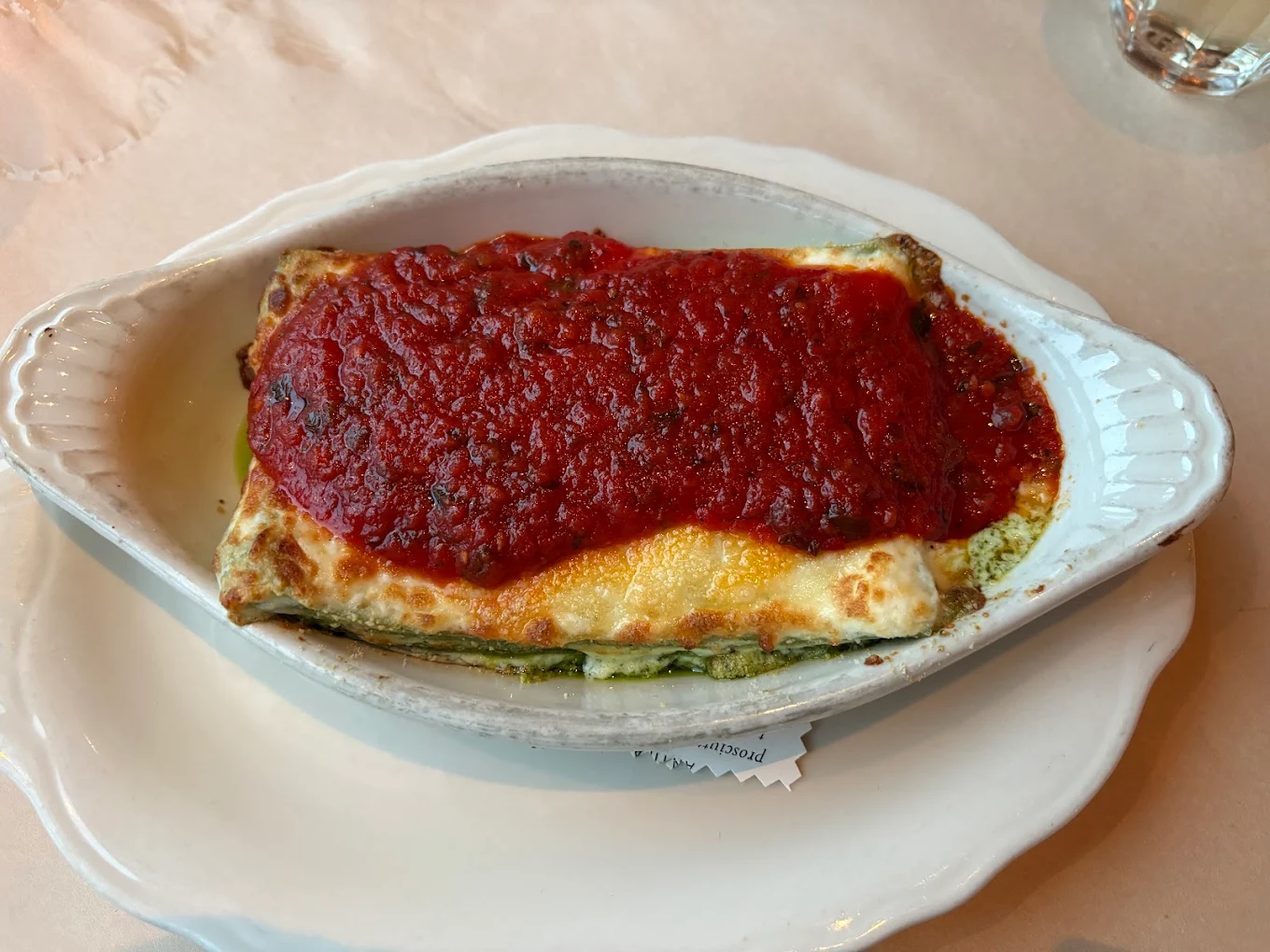 Baked lasagna topped with a thick layer of red tomato sauce, served in an oval white ceramic dish on a white plate, with part of a glass of water visible in the background.