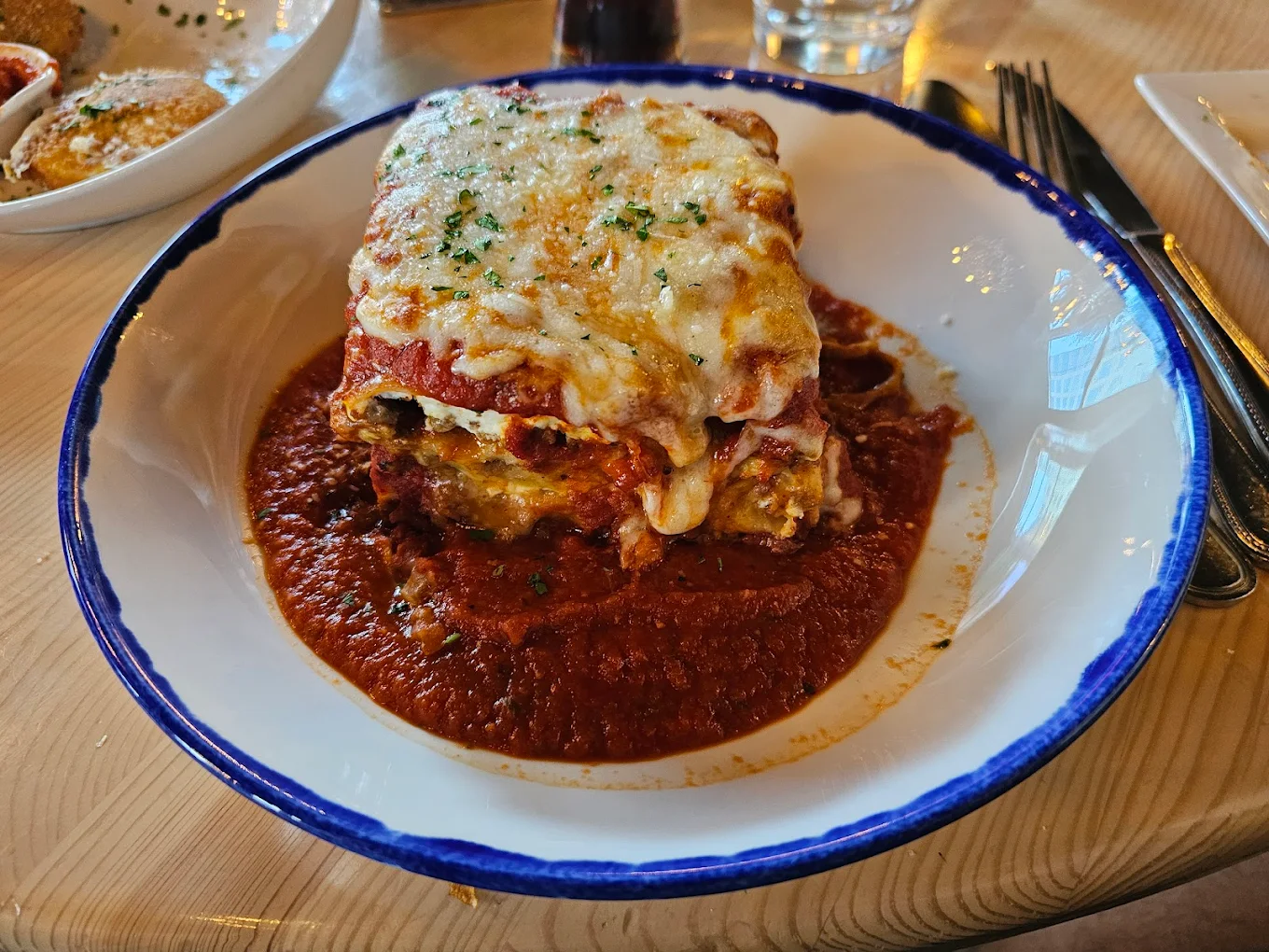 A serving of cheesy baked lasagna sits in a white bowl with a blue rim, topped with herbs and surrounded by rich tomato sauce on a wooden table.