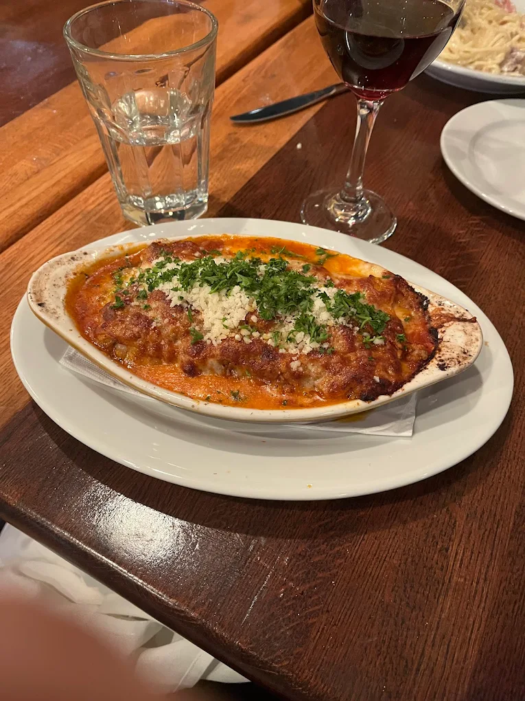 A baked dish of lasagna topped with melted cheese and fresh herbs, served on a white plate beside a glass of water and a glass of red wine on a wooden table.