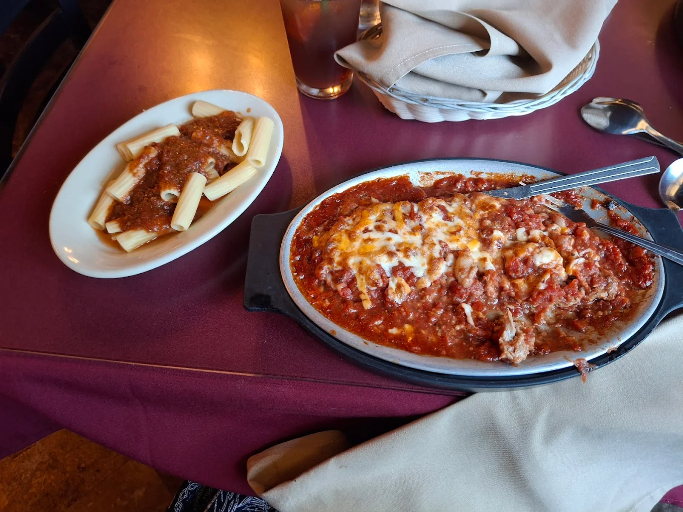 A serving dish filled with baked pasta topped with melted cheese and tomato sauce sits next to a small plate of rigatoni pasta with meat sauce on a maroon table. Silverware and a basket of napkins are nearby.