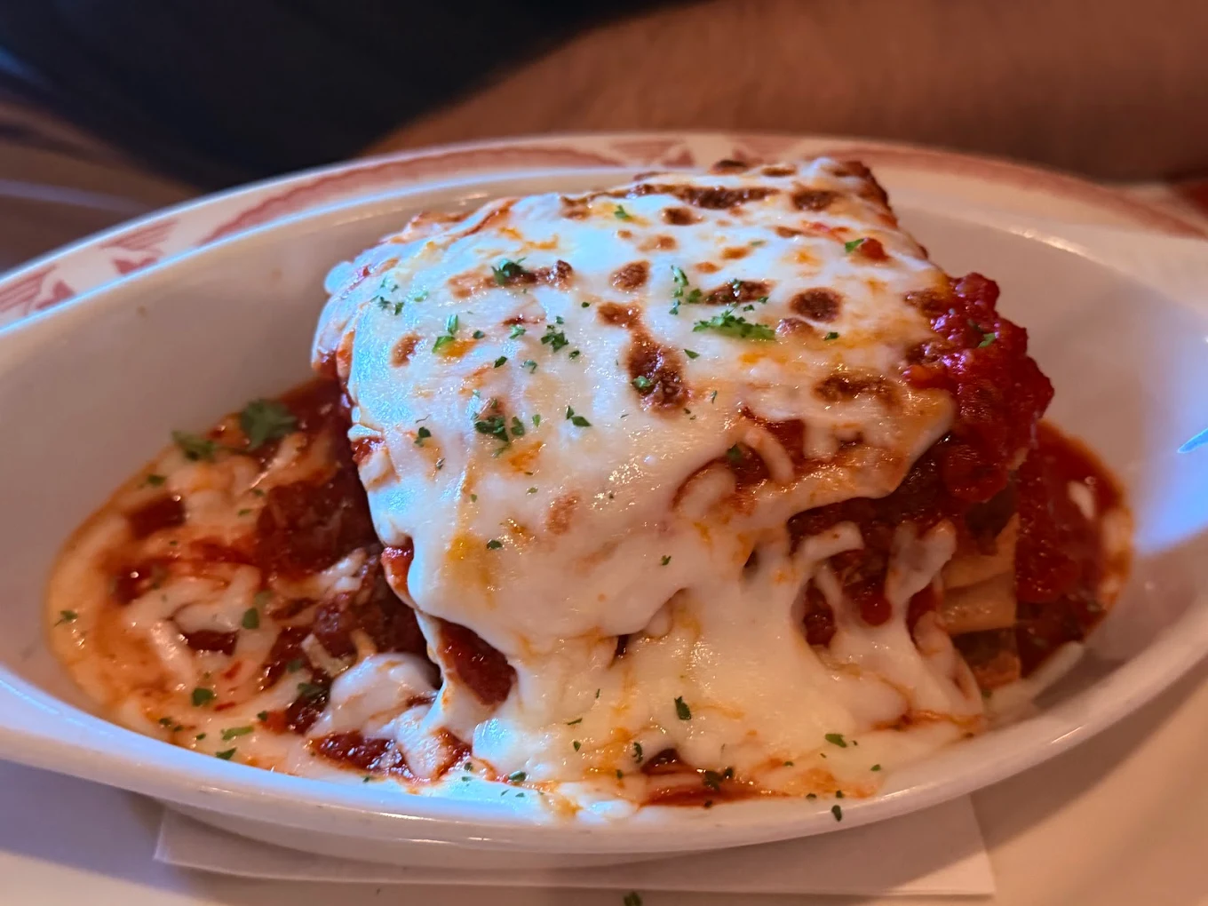 A close-up of a serving of lasagna topped with melted cheese and sprinkled with parsley, in a white dish with tomato sauce visible around the edges.