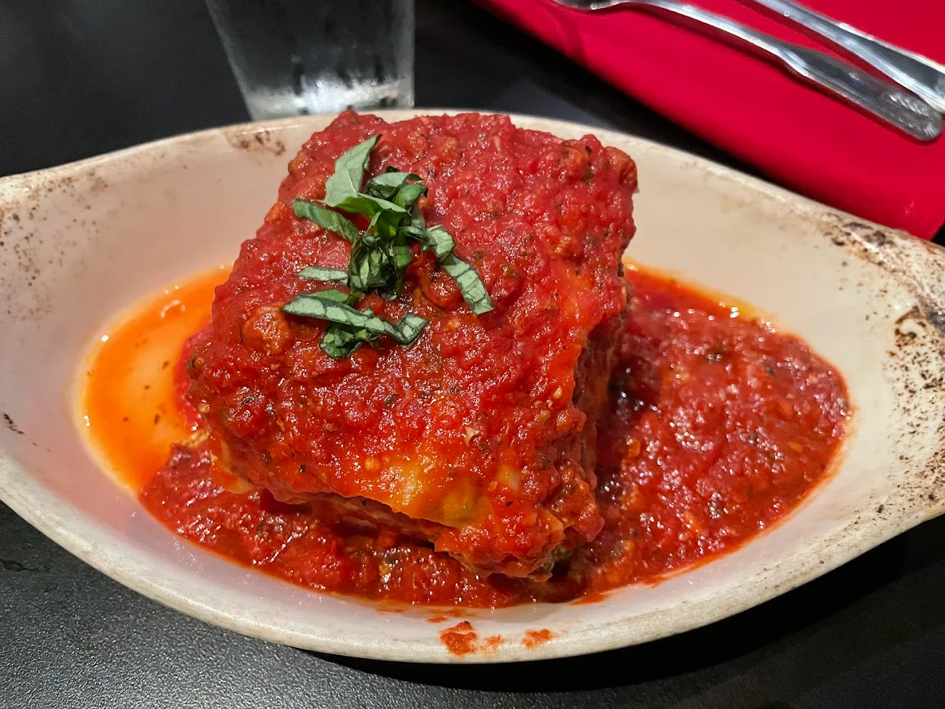 A serving of lasagna topped with rich red marinara sauce and fresh chopped basil, served in an oval dish on a dark table with a glass of water and a red napkin in the background.