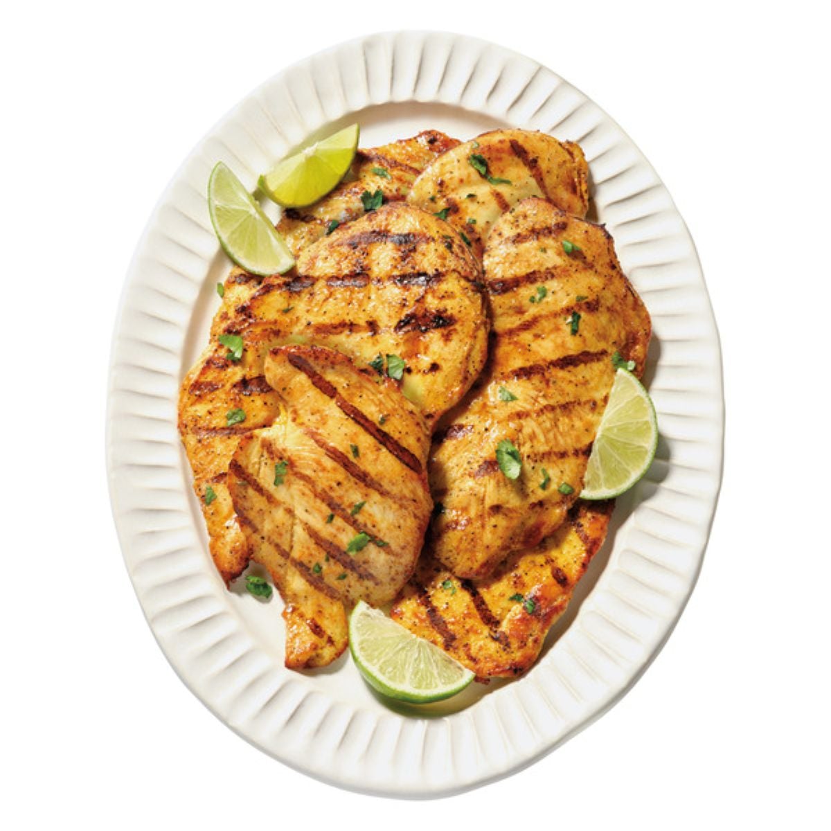 Grilled chicken breasts with grill marks are arranged on a white oval plate, garnished with chopped herbs and served with lime wedges.