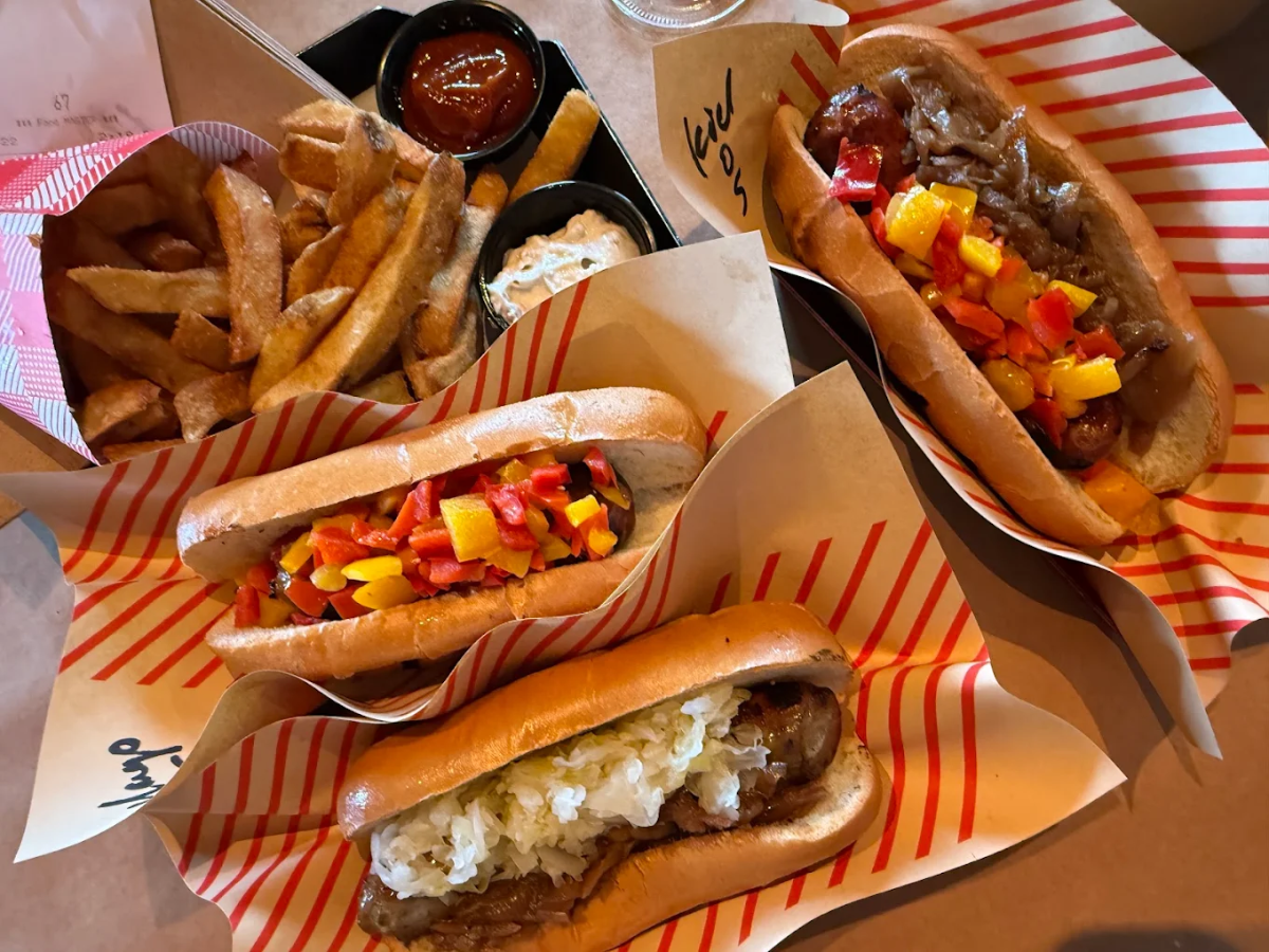 Three grilled sausages in buns with various toppings, including sautéed onions, peppers, and sauerkraut, are served on striped paper with a side of thick-cut fries, ketchup, mustard, and pickles.