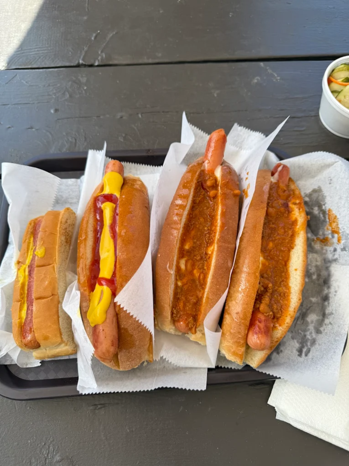 A tray with four hot dogs on white paper: two topped with mustard and ketchup, and two topped with chili. A small cup of chopped pickles or relish is visible in the top right corner.