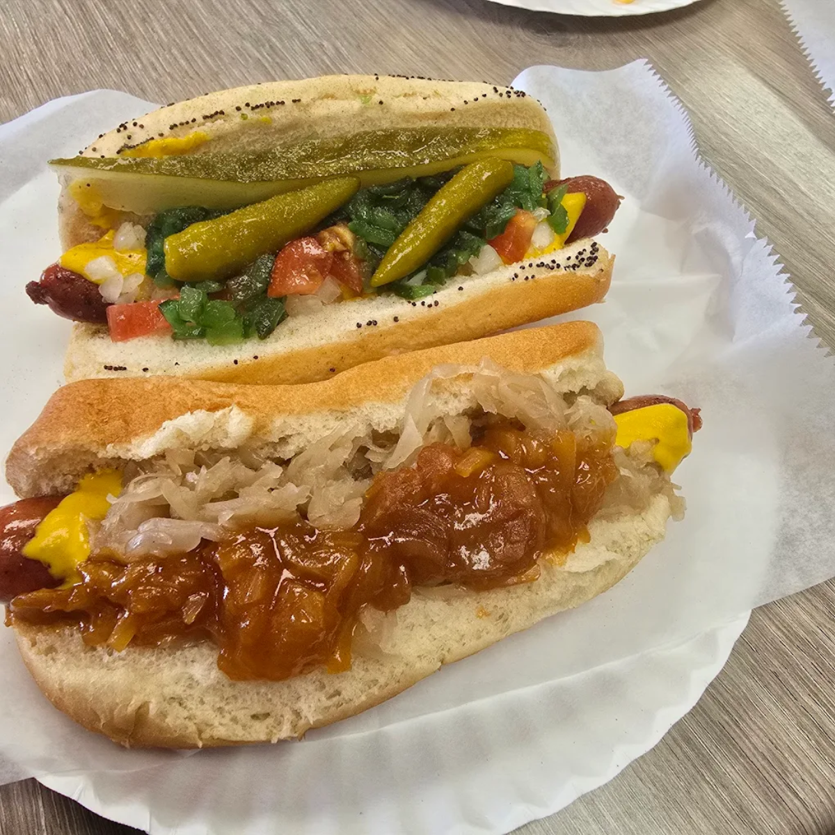 Two hot dogs on white paper plates. One is topped with pickles, tomatoes, onions, relish, mustard, and peppers; the other with sauerkraut and a stewed tomato-onion topping, both in poppy seed buns.