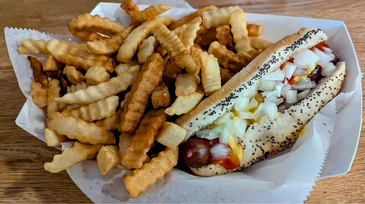 A paper tray filled with crinkle-cut French fries and a poppy seed bun hot dog topped with chopped onions, ketchup, and mustard, sitting on a wooden table.
