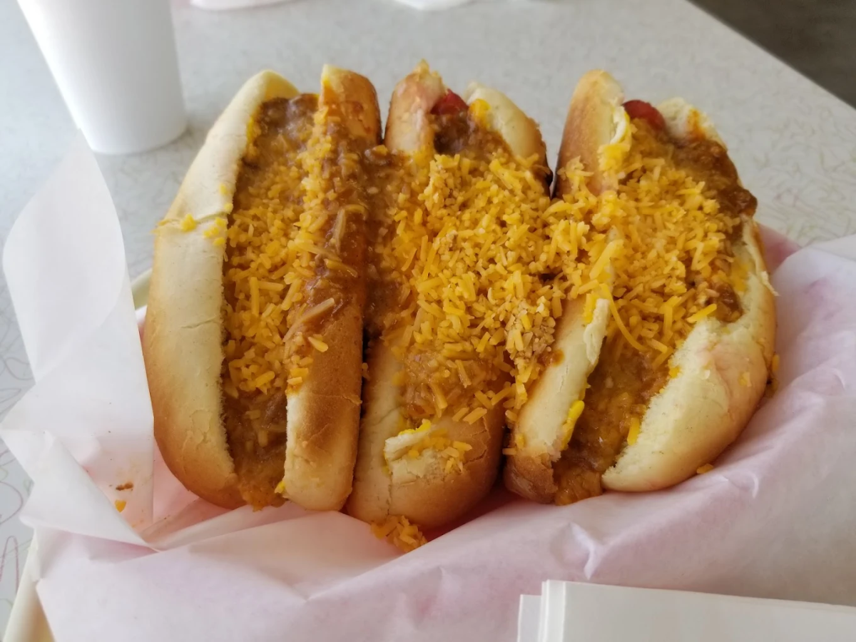 Three hot dogs in buns topped with chili and shredded cheddar cheese, served in a pink-lined basket on a light-colored table with a white cup in the background.