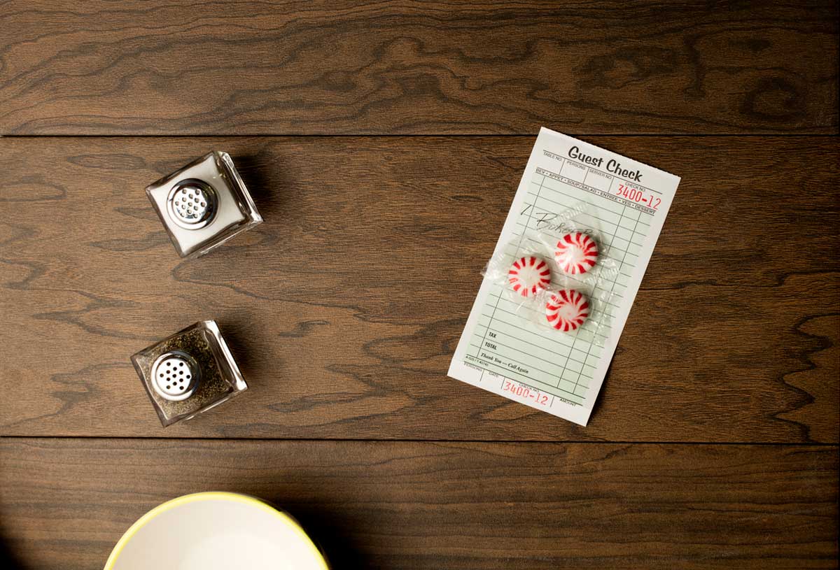 A guest check with handwriting, three steakhouse freebies like peppermint candies, salt and pepper shakers, and part of a white plate with a yellow rim on a wooden table.