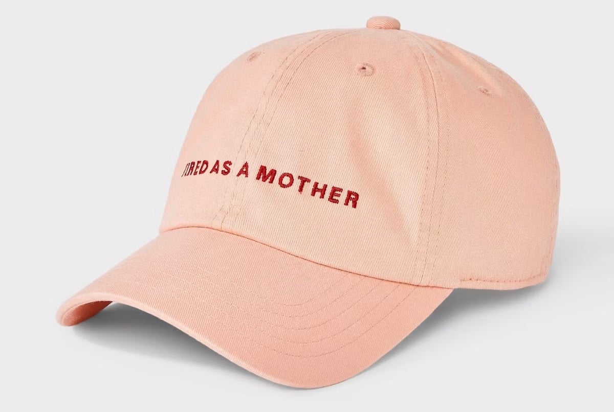 A pale pink baseball cap with the phrase "TIRED AS A MOTHER" embroidered in red on the front, shown against a plain light background—perfect for unique Mother's Day gifts.