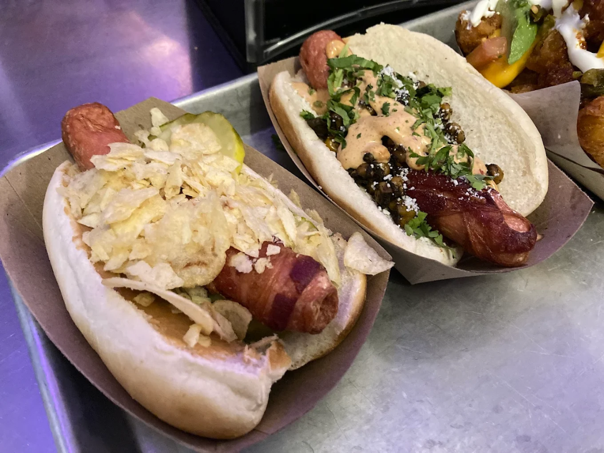 Two gourmet hot dogs in paper trays: one topped with potato chips and pickles, the other with sauce, herbs, cheese, and capers. Both hot dogs appear to be wrapped in bacon. Part of a third hot dog is visible on the side.