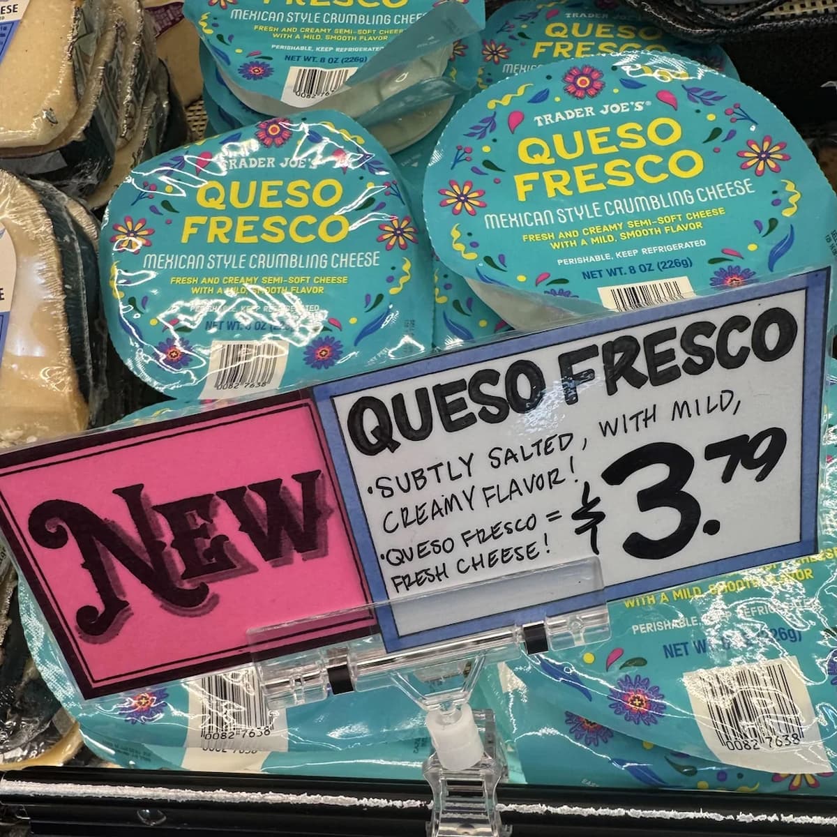 Several round packages of Trader Joe's Queso Fresco cheese are displayed behind a sign reading "NEW QUESO FRESCO, subtly salted, with mild, creamy flavor! $3.79.