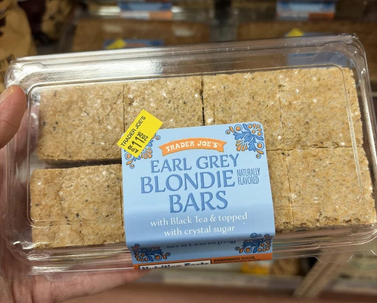 A hand holds a clear plastic container of Trader Joe's Earl Grey Blondie Bars, labeled as naturally flavored with black tea and topped with crystal sugar. Shelves with more bars are visible in the background.