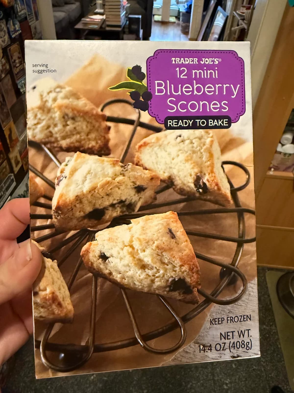 A hand holds a box of Trader Joe's 12 Mini Blueberry Scones, ready to bake. The box shows several golden scones with visible blueberries on a black wire rack. The net weight is 14.4 oz (408g).