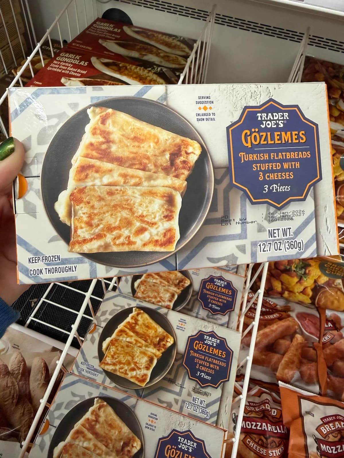 A hand holds a box of Trader Joe's Gözlemes Turkish Flatbreads stuffed with 3 cheeses in front of a freezer shelf stocked with more of the same product. The box shows an image of the cooked flatbreads on a plate.