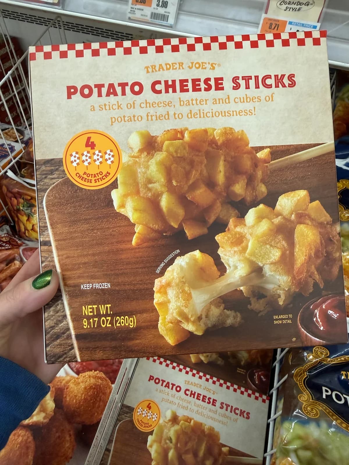 A person holding a box of Trader Joe's Potato Cheese Sticks; the packaging shows crispy, golden potato sticks filled with melted cheese. The box states “Keep Frozen” and contains 9.17 oz (260g).
