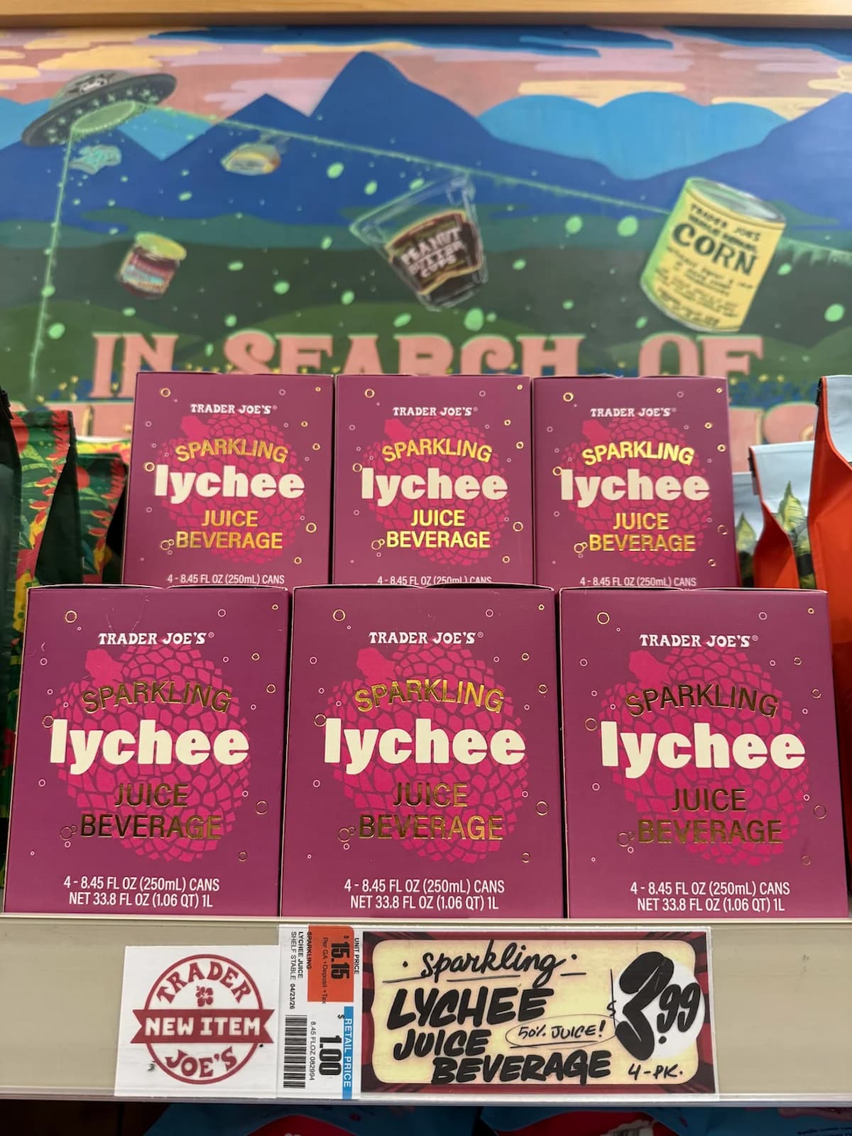 Six pink boxes of Trader Joe's Sparkling Lychee Juice Beverage are displayed on a store shelf. A sign below reads “Sparkling Lychee Juice Beverage 4-pk $3.99.” A colorful mural is in the background.