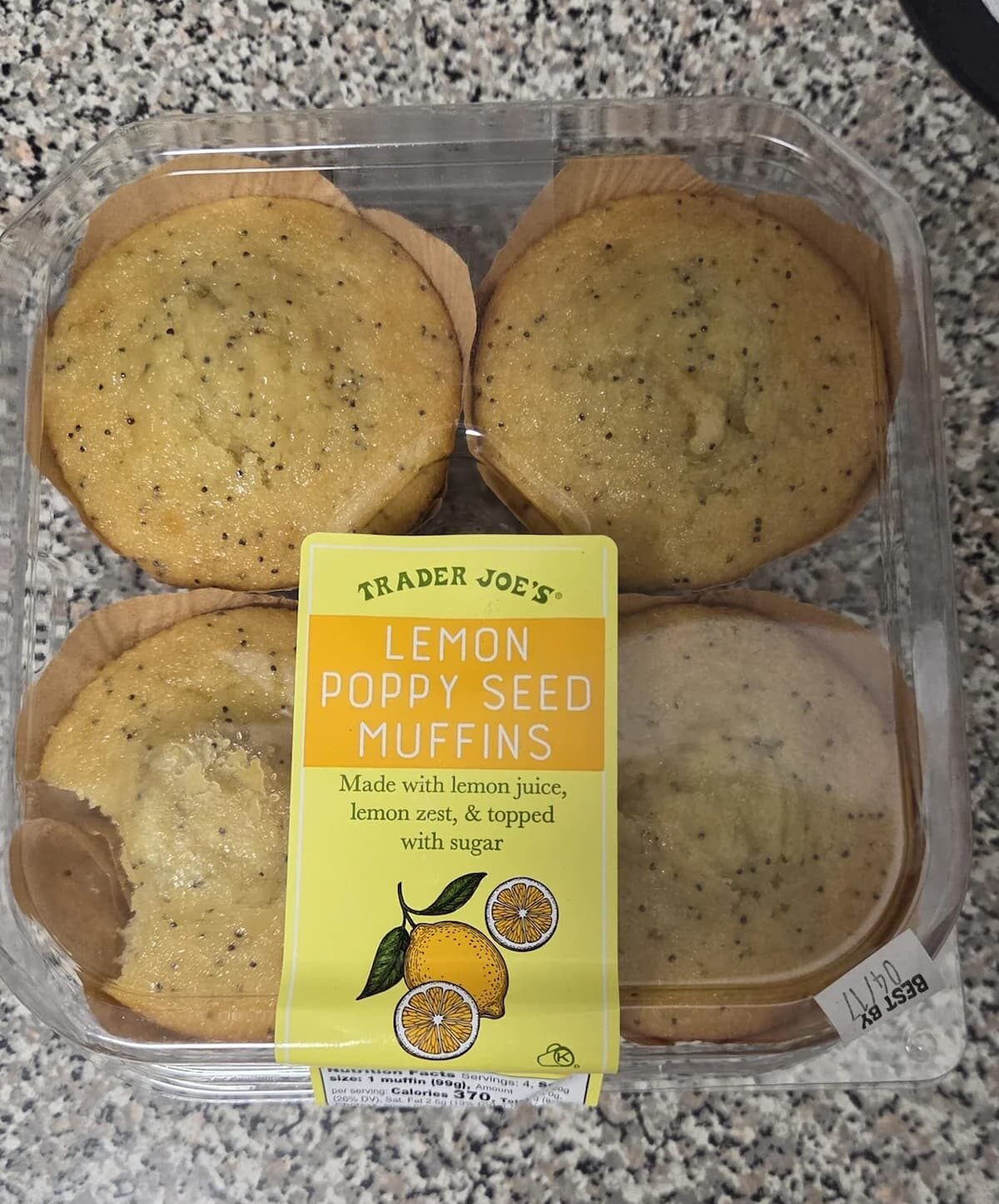 A plastic container of four Trader Joe’s lemon poppy seed muffins sits on a speckled countertop. The yellow label describes them as made with lemon juice, lemon zest, and sugar, and is decorated with lemon illustrations.