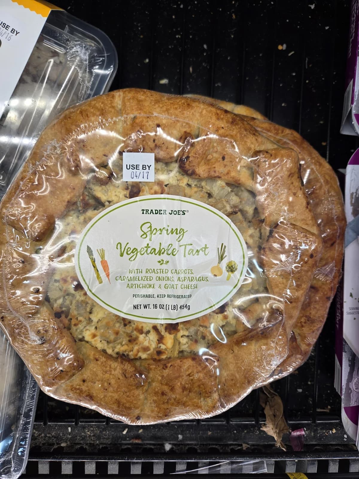 A packaged Trader Joe’s Spring Vegetable Tart with a visible label, featuring roasted carrots, caramelized onions, asparagus, artichoke, and goat cheese. The "use by" date is 04/17. The tart is on a store shelf.