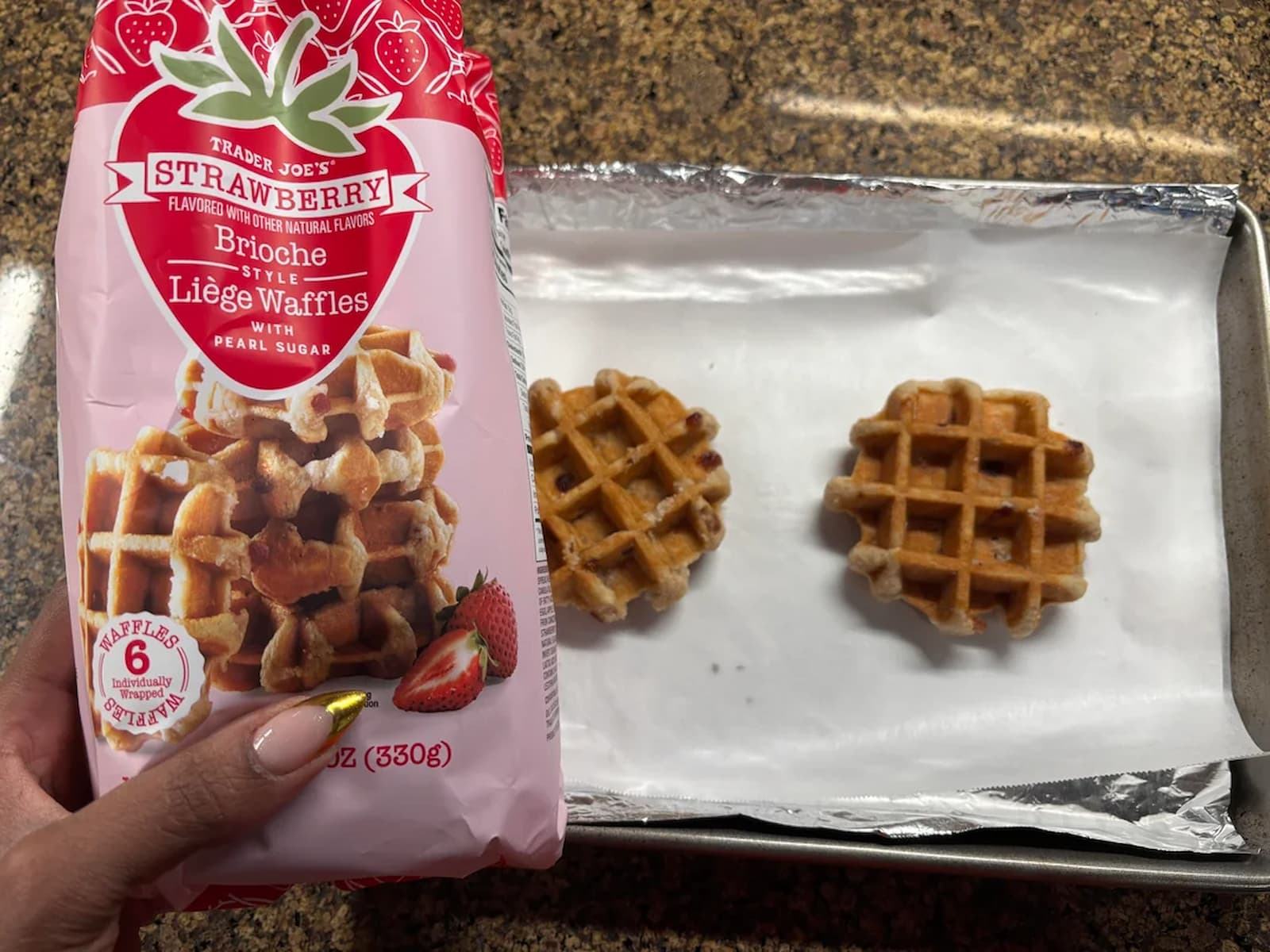 A hand holds a package of Trader Joe’s Strawberry Brioche Liège Waffles over a baking sheet with two small waffles spaced apart on parchment paper. The waffles are golden brown and the counter is granite.