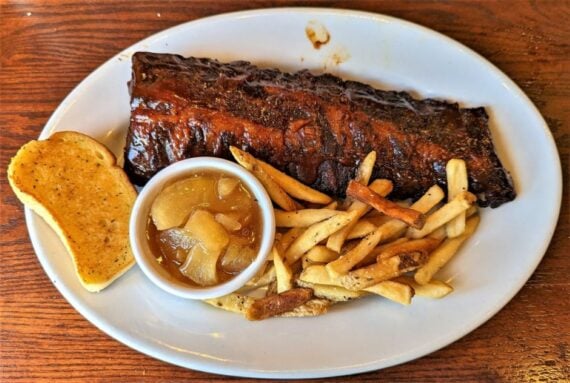 A plate with a rack of glazed ribs, a serving of French fries, a slice of garlic bread, and a small bowl of cooked apples, all on a wooden table.
