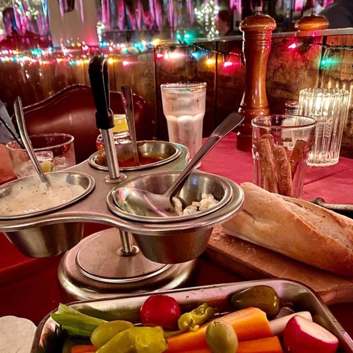 A cozy restaurant table with colorful string lights, a tray of fresh vegetables, a baguette, breadsticks, and a three-bowl condiment server. Glasses, candles, and a pepper grinder sit on the red tablecloth.