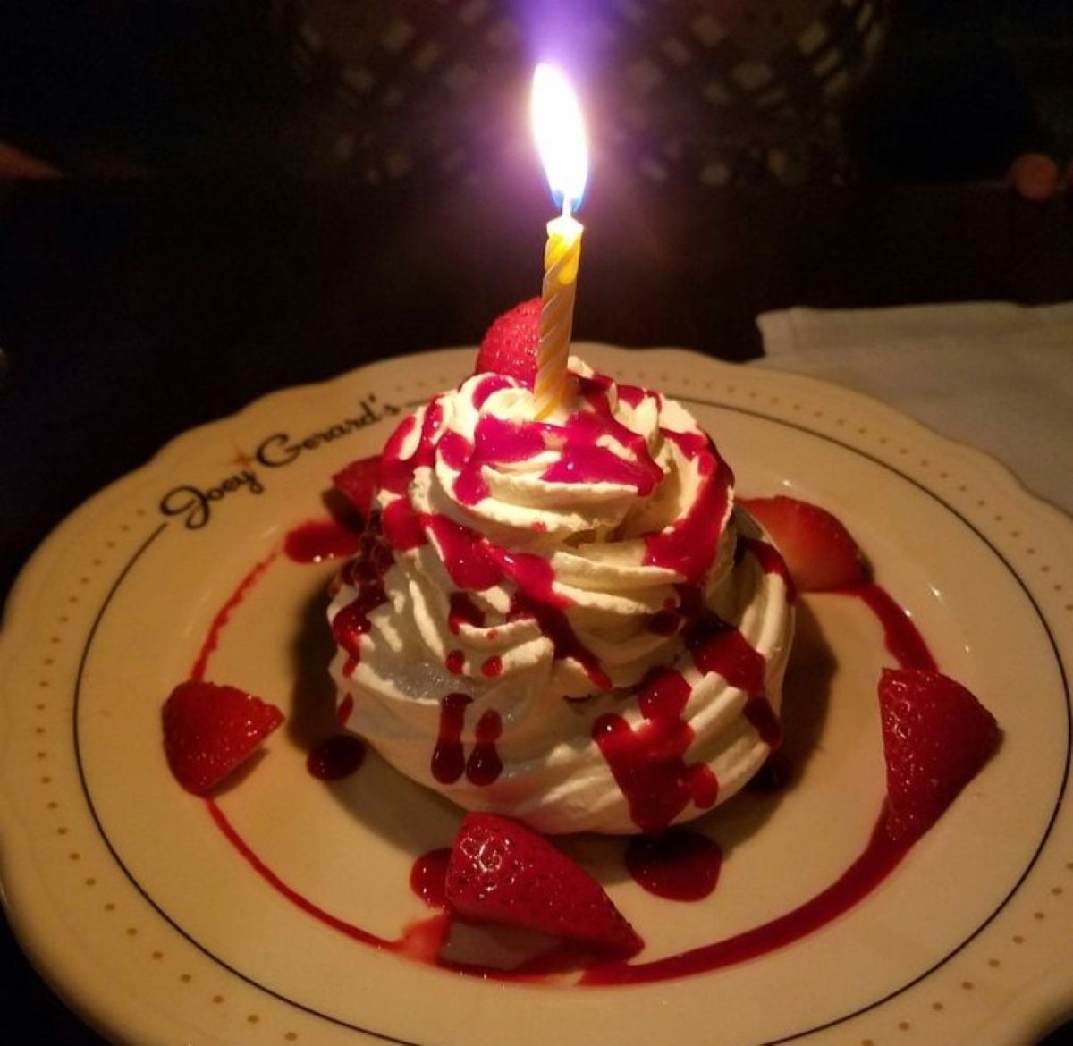 A dessert topped with whipped cream, strawberry sauce, and a lit birthday candle sits on a plate, garnished with fresh strawberry pieces.