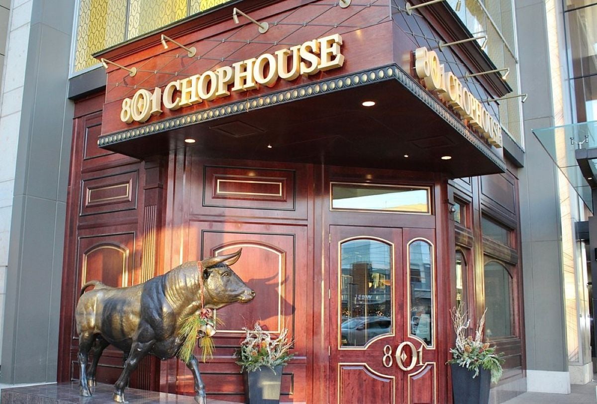 The entrance of 801 Chophouse features dark wood-paneled doors, gold signage, decorative plants, and a large bronze bull statue beside the doorway—showing no signs of bankruptcy here.