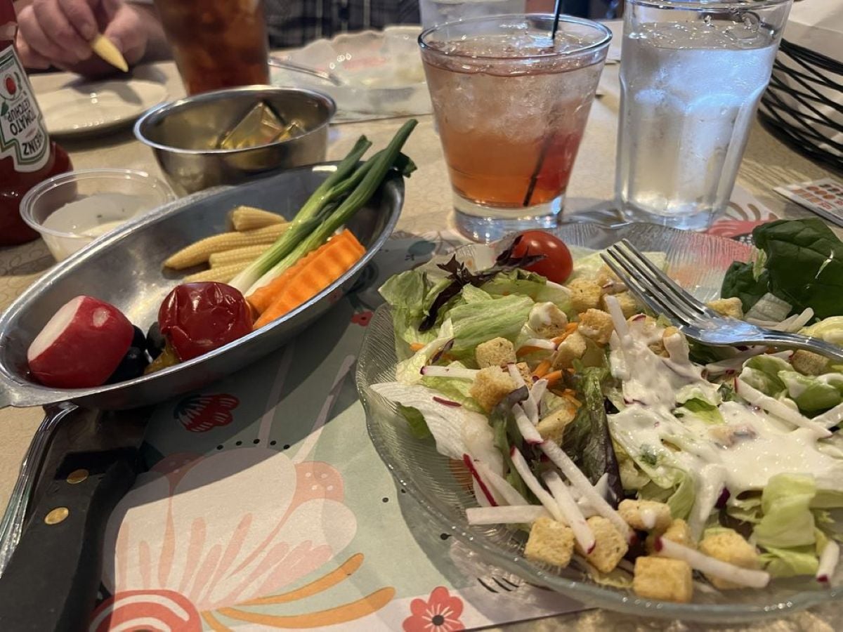 A table with a fresh green salad topped with croutons and ranch dressing, a plate of raw vegetables, a glass of iced water, and a red drink, with condiments and a bread basket in the background.