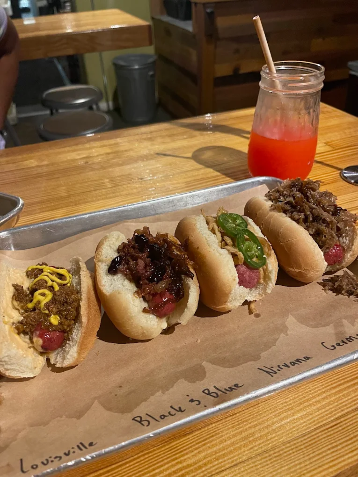 A tray with four gourmet hot dogs, each with different toppings, sits on a wooden table next to a mason jar filled with a red drink. The tray's paper liner is labeled with the names of each hot dog variety.
