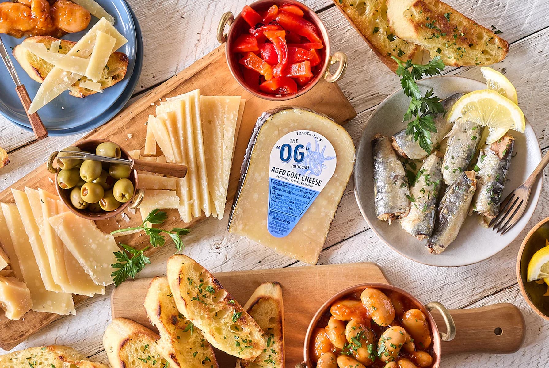 A charcuterie spread with aged goat cheese, toasted bread slices, olives, roasted red peppers, sardines, marinated beans, and cheese wedges on wooden boards and plates, garnished with parsley.