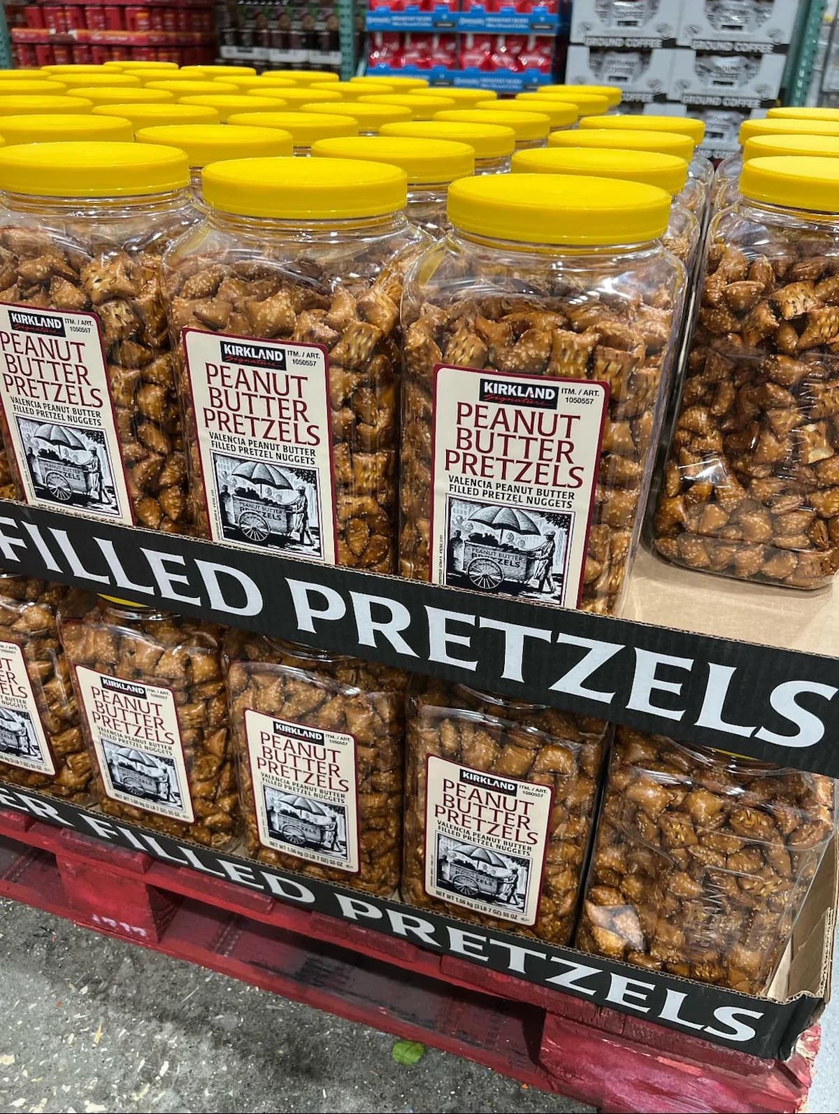 Several large plastic jars with yellow lids, filled with Kirkland brand peanut butter pretzel nuggets, are stacked on a shelf in a store. The jars display prominent labels and are arranged in two rows.