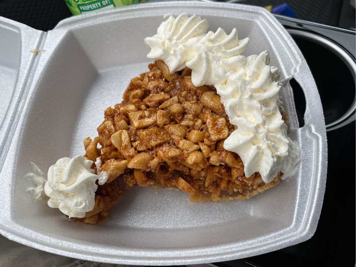 A slice of nut pie with a golden-brown filling and a crust, topped with swirls of whipped cream, served in a white takeout container. Part of the slice has been eaten.