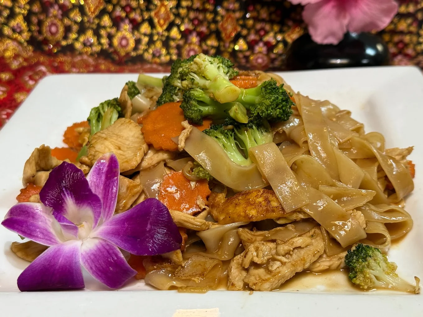 A plate of stir-fried flat rice noodles with chicken, broccoli, carrots, and tofu is garnished with a purple orchid flower. A colorful, patterned cloth and a pink flower are in the background.