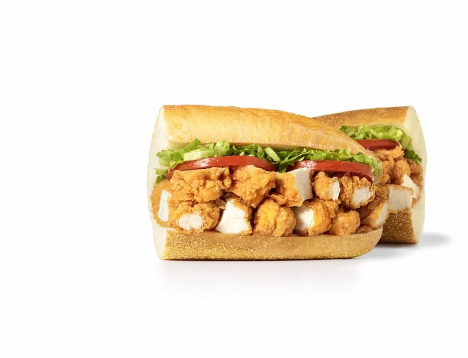 A sandwich with crispy fried chicken pieces, sliced tomatoes, and shredded lettuce on a sliced baguette, set against a white background.