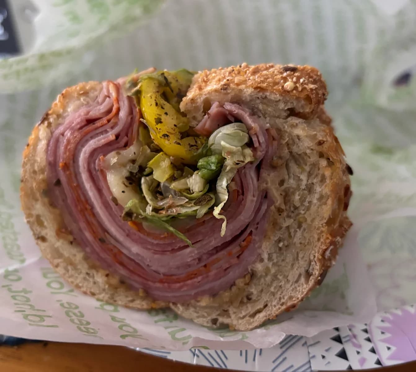 A close-up of a sandwich with multiple layers of sliced deli meats, shredded lettuce, yellow peppers, and herbs on multigrain bread, partially wrapped in printed sandwich paper.