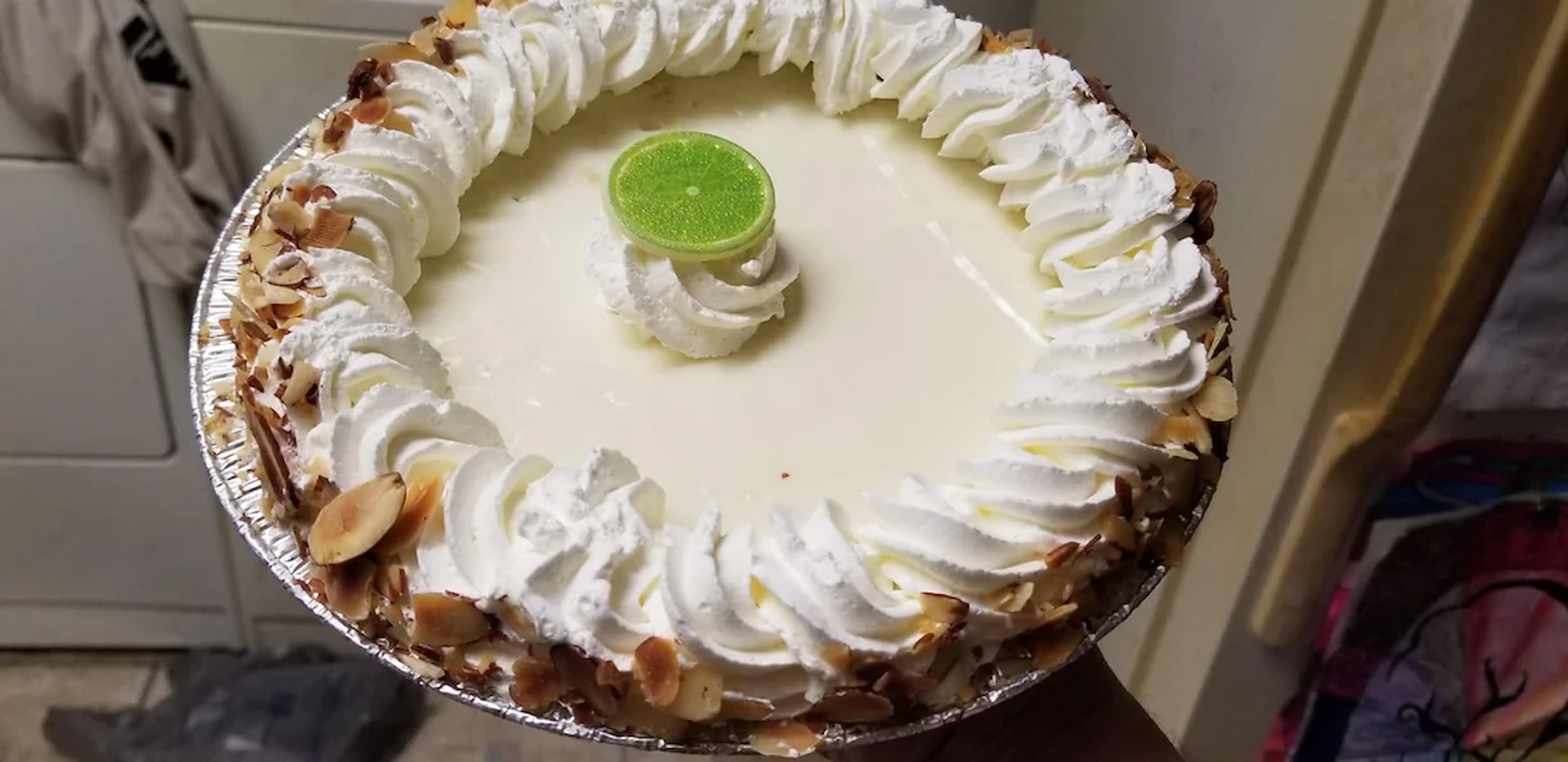 A key lime pie with whipped cream swirls around the edges, topped with a slice of lime in the center, and garnished with toasted almond slices on the crust.
