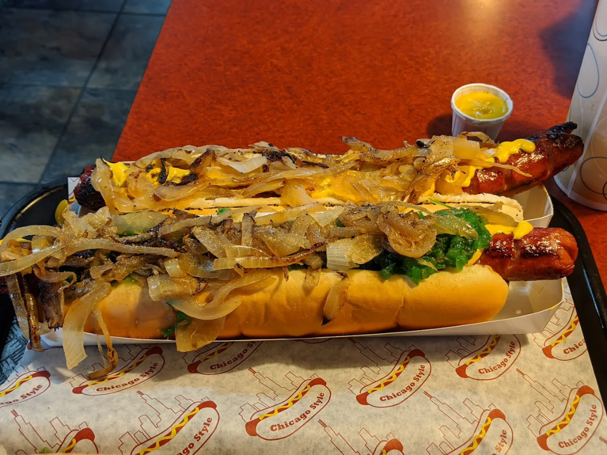 Two grilled hot dogs topped with grilled onions, mustard, and relish, served in buns on a tray with a side of mustard, on a red table.