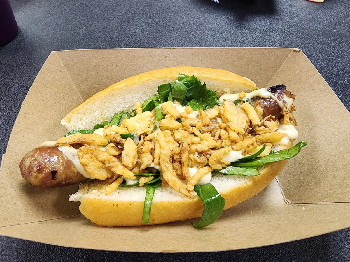 A sausage in a bun topped with fresh spinach, crispy fried onions, and a drizzle of sauce, served in a brown paper tray on a dark surface.