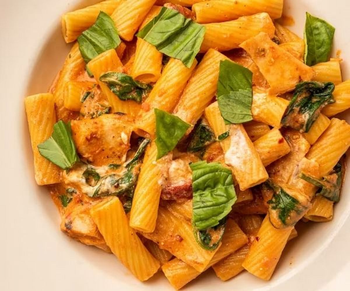 A close-up of rigatoni pasta in a creamy tomato sauce, mixed with spinach and topped with fresh basil leaves—just like the popular dishes at Italian chain restaurants in America—served on a white plate.