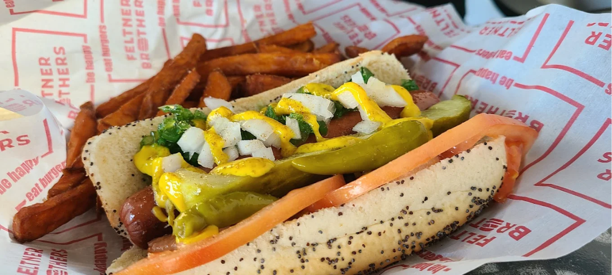A Chicago-style hot dog in a poppy seed bun topped with mustard, onions, relish, pickle spears, tomato slices, and sport peppers, served with a side of sweet potato fries on branded paper.