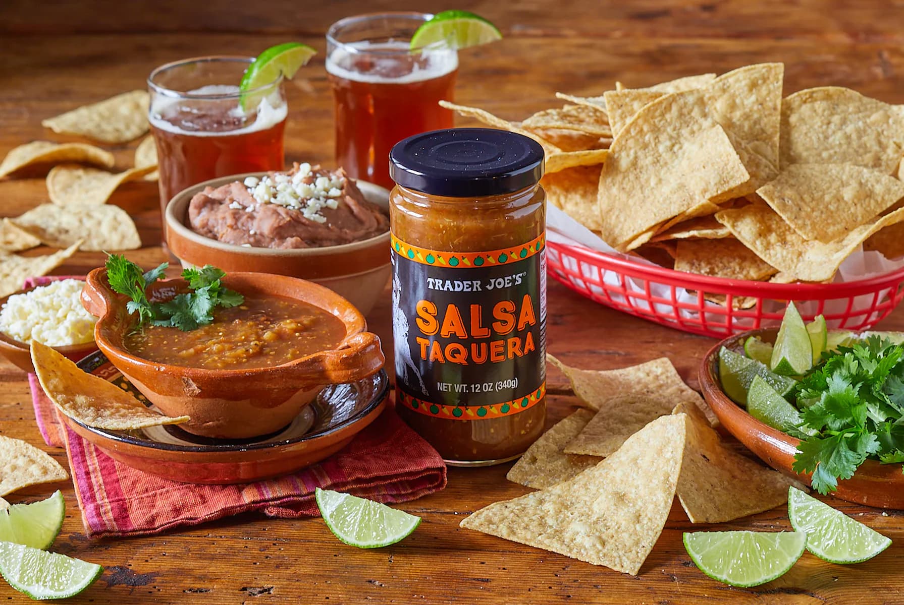 A jar of Trader Joe’s Salsa Taquera sits on a wooden table with tortilla chips, salsa in a clay bowl, refried beans, lime wedges, fresh cilantro, and glasses of amber drinks in the background.