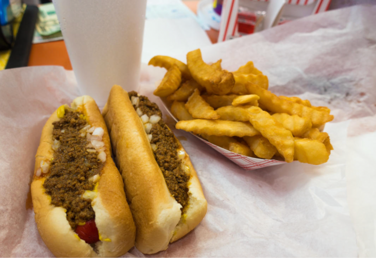 Two chili dogs topped with onions and mustard on a tray, served with a portion of crinkle-cut French fries and a large white cup, set on a table covered with parchment paper.