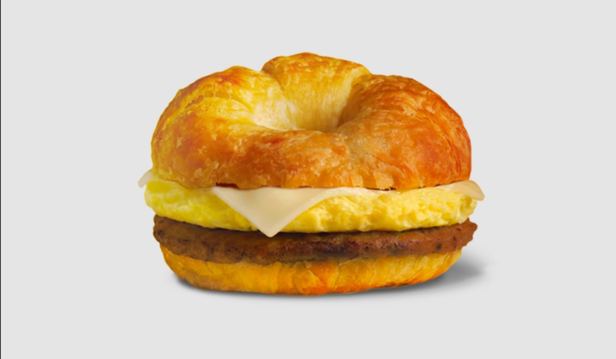 A breakfast sandwich with a croissant bun, egg, sausage patty, and a slice of cheese, set against a plain light background.