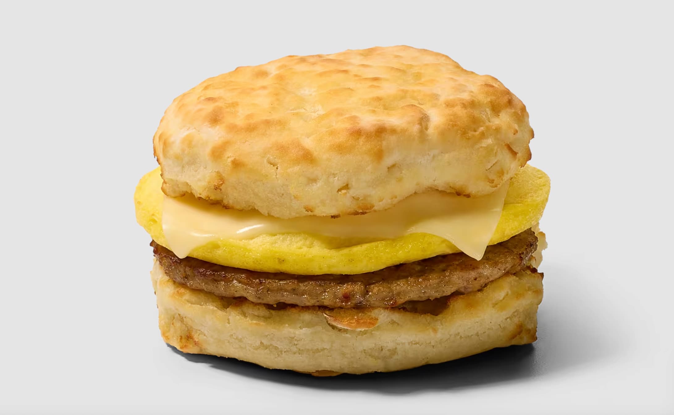 A breakfast sandwich with a fluffy biscuit, folded egg, melted cheese, and a sausage patty on a plain light gray background.