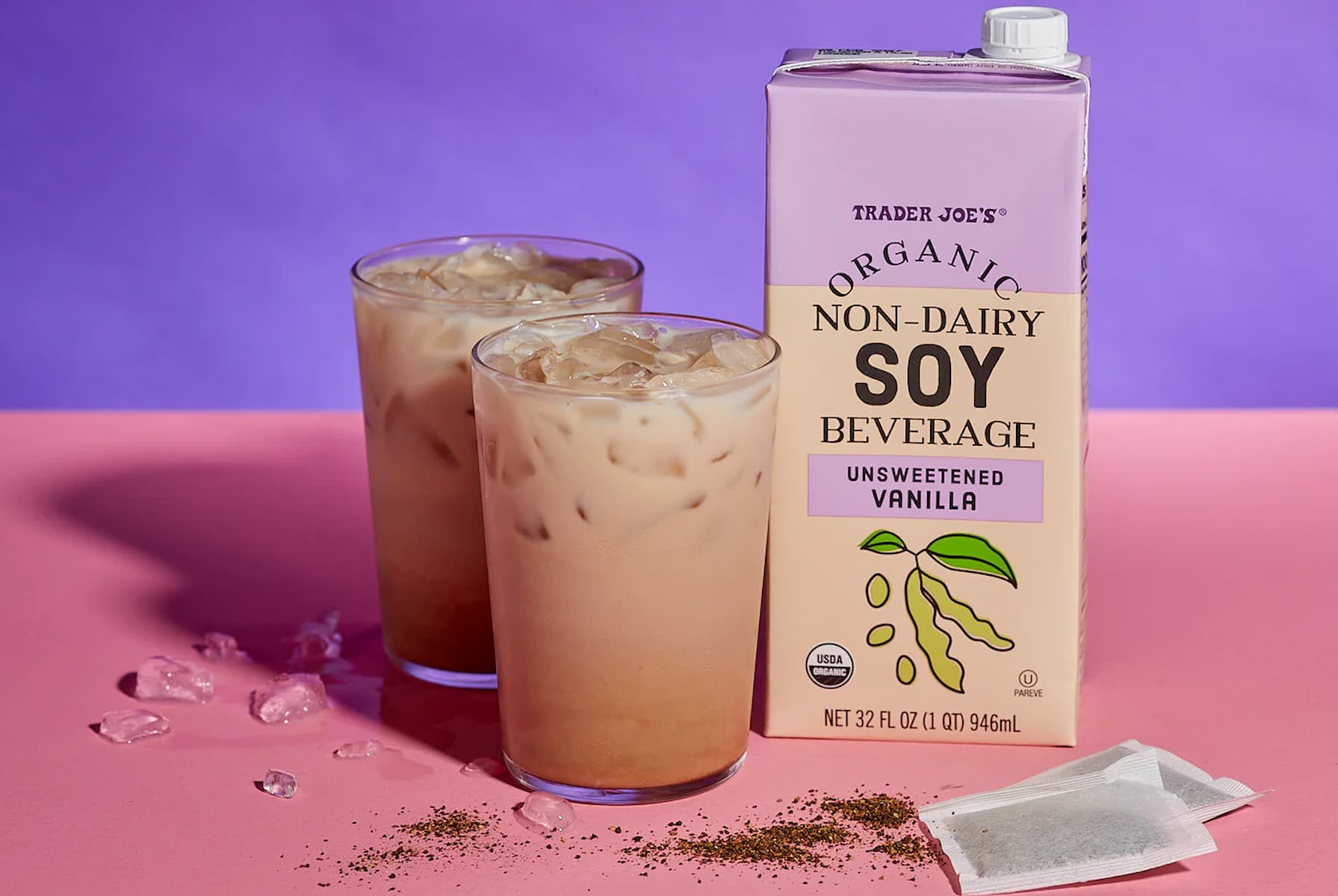 Two glasses of iced latte sit next to a carton of Trader Joe's Organic Non-Dairy Soy Beverage in Unsweetened Vanilla flavor. One tea bag, some loose tea leaves, and scattered ice are on a pink surface with a purple background.