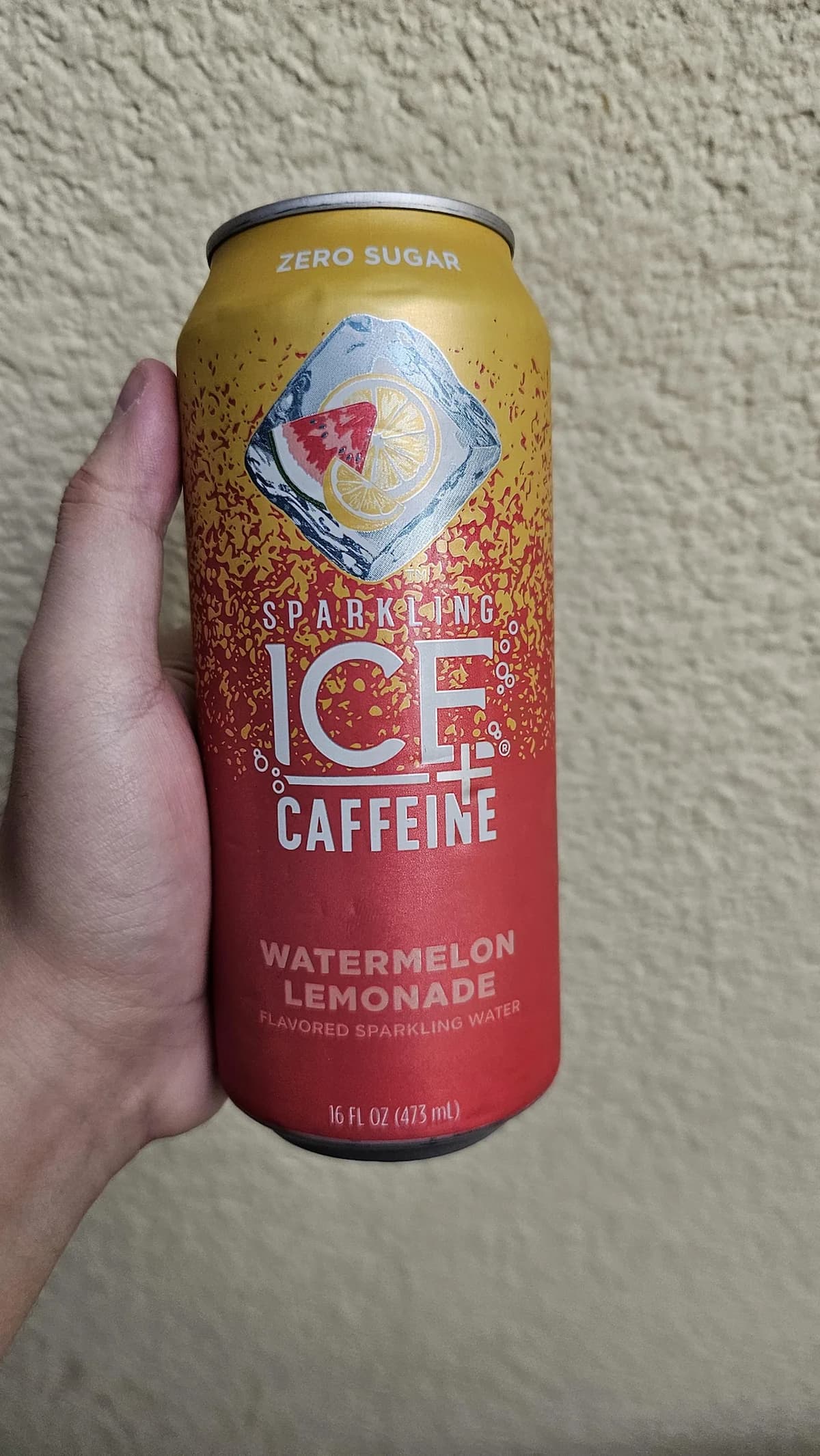 A hand holds a 16 fl oz can of Sparkling Ice + Caffeine in Watermelon Lemonade flavor. The can is red and yellow with "Zero Sugar" and a lemon slice logo on top. The background is a textured beige wall.